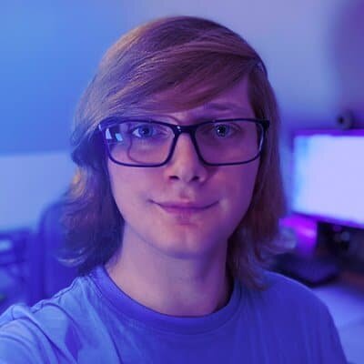 Headshot of Adam Greenough, a young adult male with shoulder length blonde hair and glasses in front of a computer desk